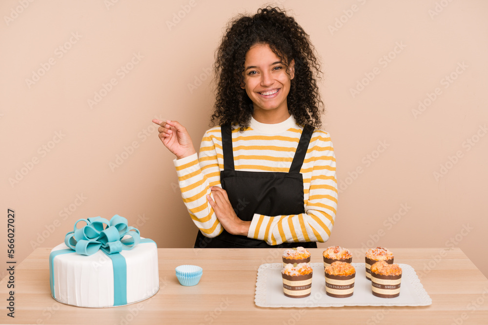 Young african american woman preparing a sweet cake and muffins on a table smiling cheerfully pointing with forefinger away. Young african american woman preparing a sweet cake and muffins on a table smiling cheerfully pointing with forefinger away.
