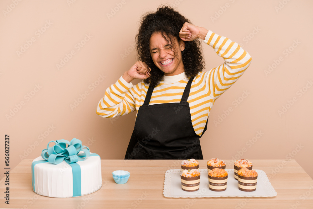 Young african american woman preparing a sweet cake and muffins on a table celebrating a special day, jumps and raise arms with energy. Young african american woman preparing a sweet cake and muffins on a table celebrating a special day, jumps and raise arms with energy.