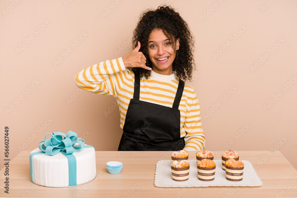Young african american woman preparing a sweet cake and muffins on a table showing a mobile phone call gesture with fingers. Young african american woman preparing a sweet cake and muffins on a table showing a mobile phone call gesture with fingers.