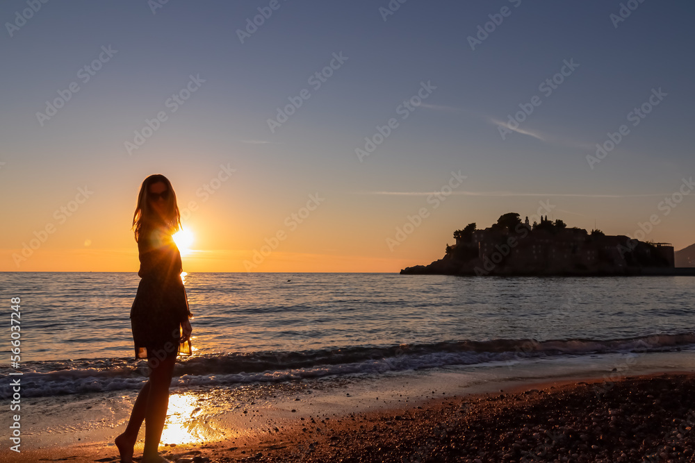 Silhouette of barefoot woman on sand beach with scenic romantic view on idyllic island Sveti Stefan at sunset, Budva Riviera, Adriatic Mediterranean Sea, Montenegro, Europe. Summer vacation at seaside