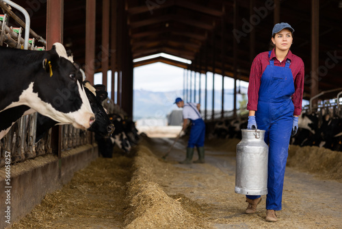 Portraif of young caucasian woman dairy farm worker carrying metal milk can in cowshed