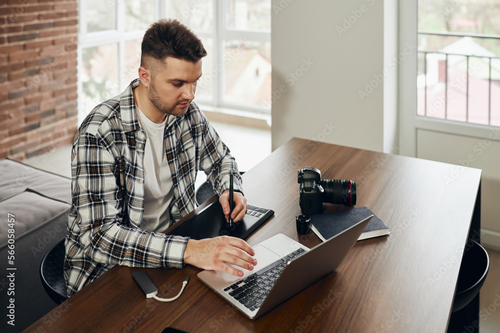 A professional photographer is sitting at his office desk. Man in office with digital graphics tablet and pen for drawing. A camera, a laptop, a card reader are all around on the table.