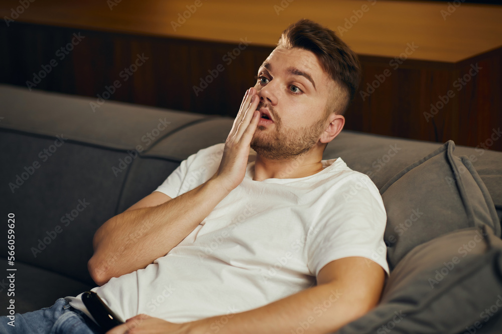 Fototapeta premium A young man in a white T-shirt, exhausted, fell asleep in front of the TV. Lazy man watching television at night alone.