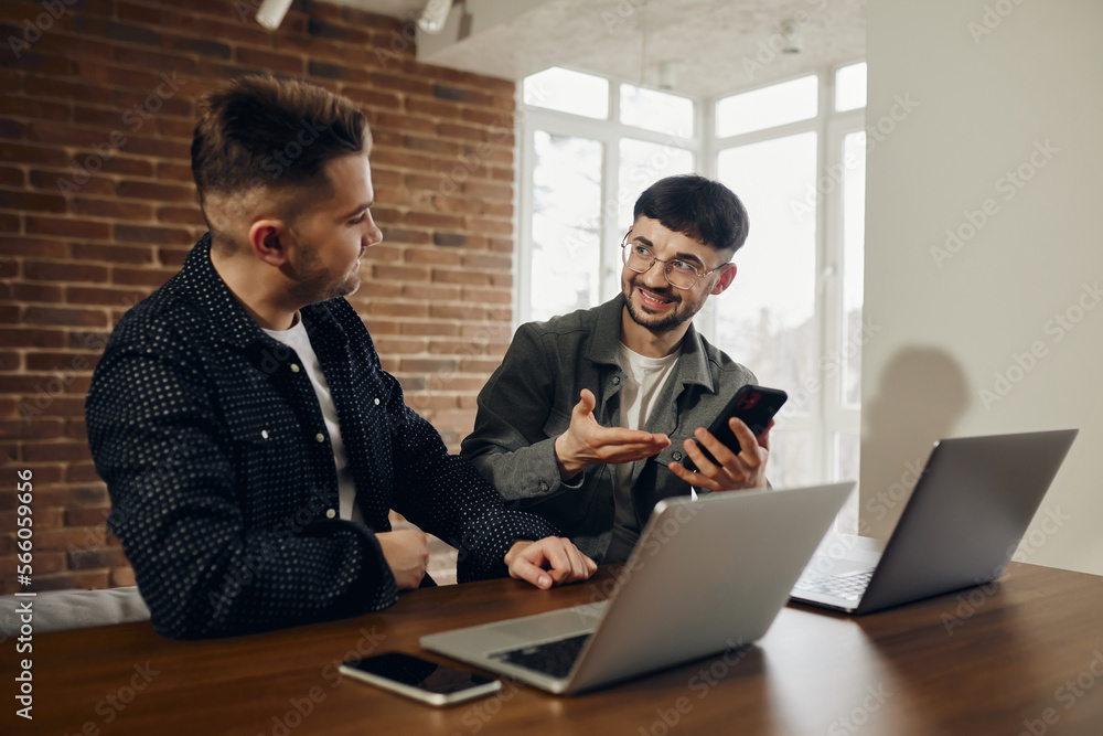 Two happy young men are working in the office on laptops. One shows the other something on the phone. Two happy young men are working in the office on laptops. One shows the other something on the phone.