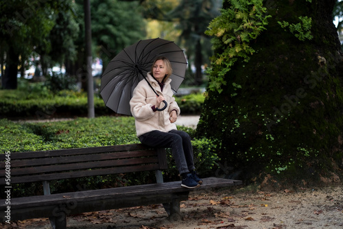 A woman with an umbrella sits on the back of a bench in the Park.