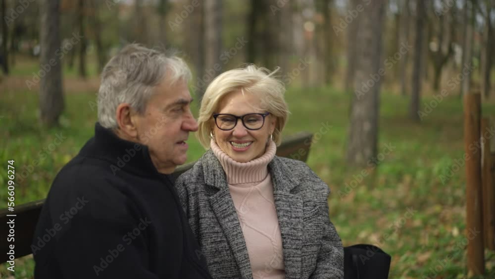 Senior couple sitting on bench while resting during a walk, embracing and discussing beautiful summer nature in the forest Senior couple sitting on bench while resting during a walk, embracing and discussing beautiful summer nature in the forest