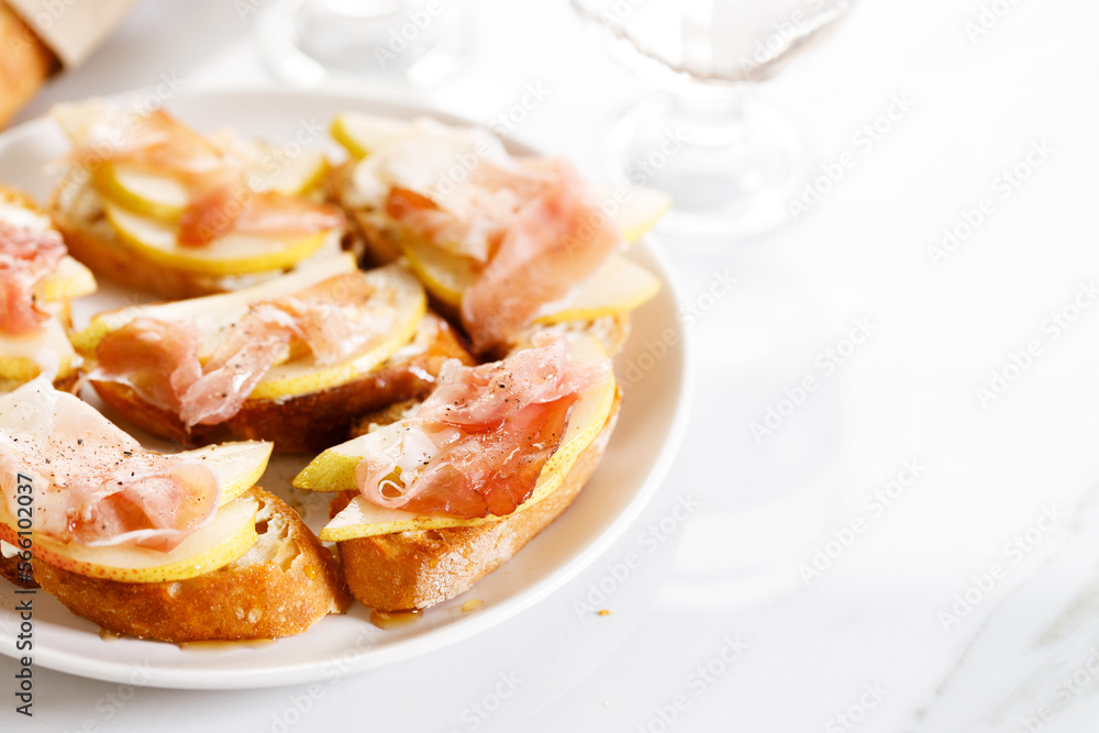 Crostini with cheese, sliced pear and prosciutto on white plate.