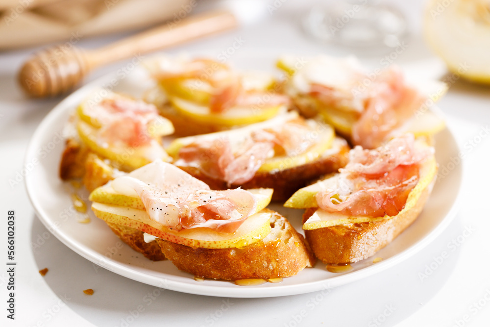 Crostini with cheese, sliced pear and prosciutto on white plate.