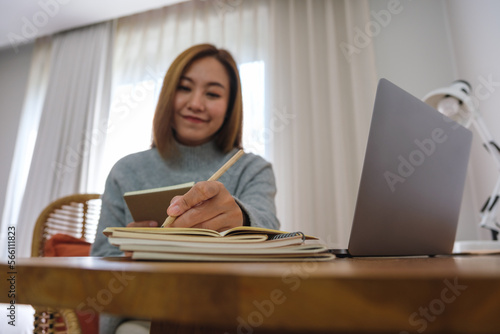 Portrait image of a young woman writing on notebook while working or study on laptop computer at home © Farknot Architect