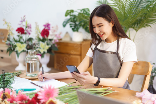 Flower shop concept, Female florist checking flower orders on smartphone and take notes in notebook