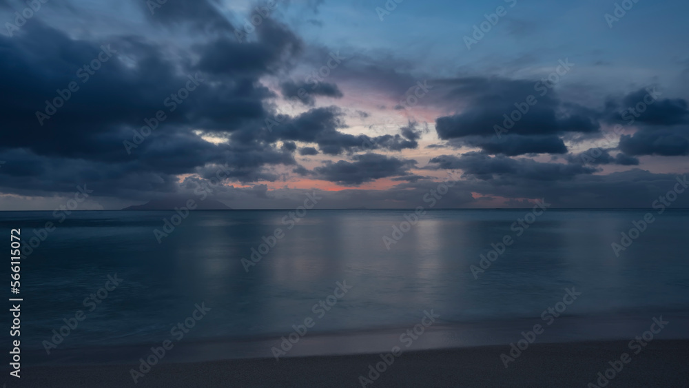 Evening twilight on a sandy tropical beach. The clouds in the sky are highlighted in pink. The outline of the island on the horizon. Reflection on the ocean surface. Seychelles. Mahe. Beau Vallon.
