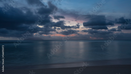 Evening twilight on a sandy tropical beach. The clouds in the sky are highlighted in pink. The outline of the island on the horizon. Reflection on the ocean surface. Seychelles. Mahe. Beau Vallon. 