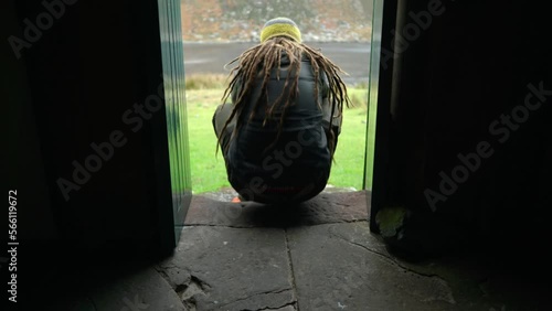A man with dreadlocks and wearing hiking gear walks into shot to sit on a step in a narrow doorway of a bothy in the Highlands of Scotland to enjoy the view outdoors. photo