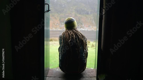 A man with dreadlocks and wearing hiking gear sits on a step in a narrow doorway of a bothy in the Highlands of Scotland to enjoy the view outdoors. photo