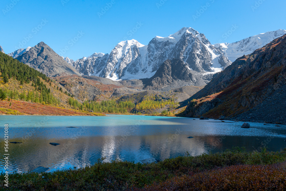 The shore of the alpine lake Shavlinskoye in Altai in the shade against the backdrop of sun-drenched peaks with snow and glaciers. Mountains Dream, Fairy Tale, Beauty.