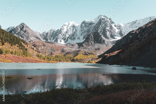 The shore of the alpine lake Shavlinskoye in Altai in the shade against the backdrop of sun-drenched peaks with snow and glaciers.