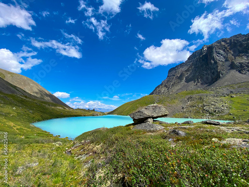 Turquoise water of Karakabak lake in Altai mountains with mountain peaks with under clouds and green grass with hanging stone.
