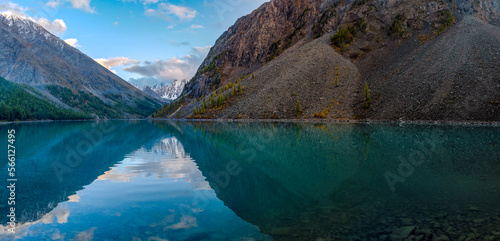 Panorama of the turquoise lake Shavlinskoe in the shade with stones among the mountains with reflection of the peaks with glaciers and snow in Altai.
