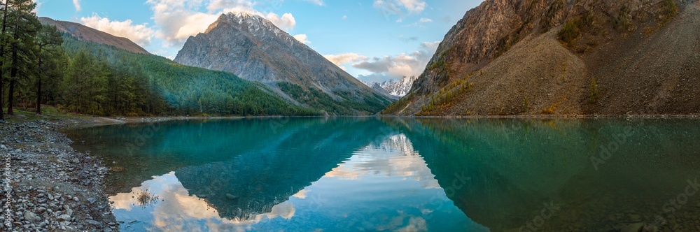 Panorama of Lake Shavlinskoe in the shade with stones among mountains with reflection of peaks with glaciers and snow in Altai in the evening in autumn.