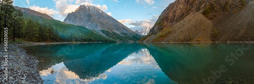 Panorama of Lake Shavlinskoe in the shade with stones among mountains with reflection of peaks with glaciers and snow in Altai in the evening in autumn.