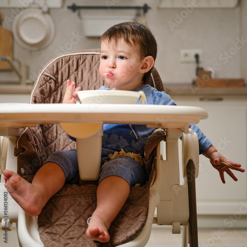 A funny child is eating a grated apple with his mouth full while sitting on a kitchen chair. Hungry baby boy shoves food in his mouth, humor. Kid aged one year four months photo