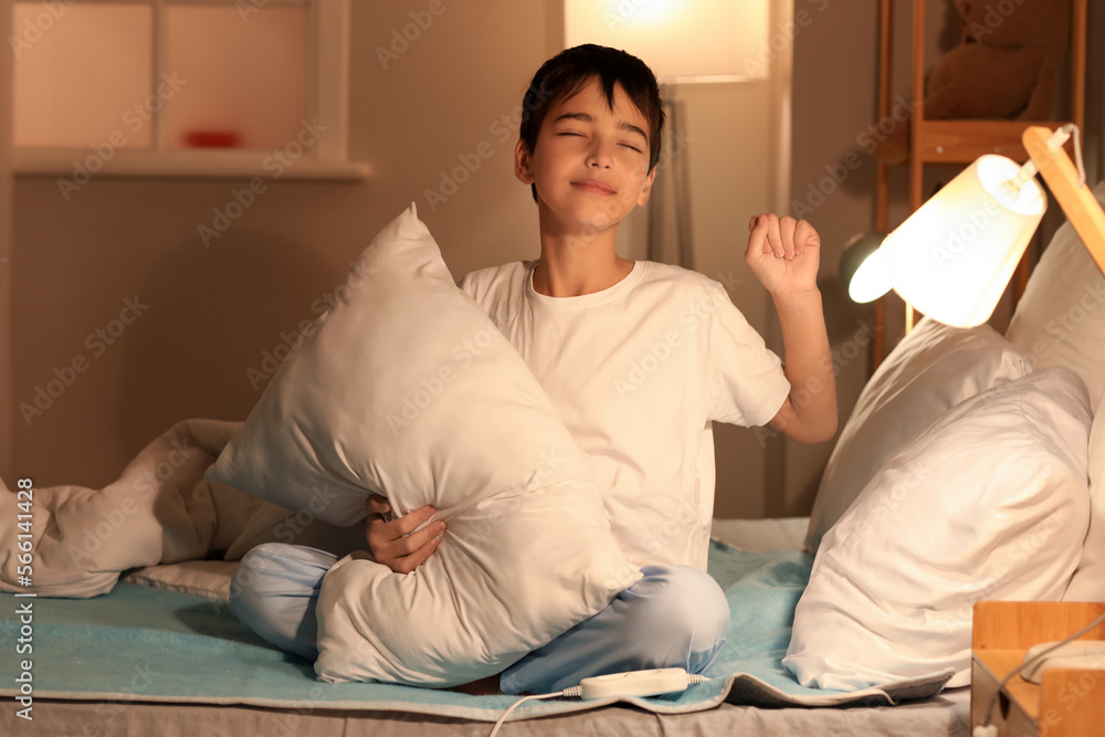 Fototapeta premium Sleepy little boy with pillow sitting on electric heating pad in bedroom at night