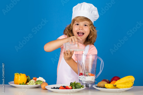Child chef cook prepares milk shake smoothie in isolated blue studio background. Kids cooking. Teen boy with apron and chef hat preparing a healthy vegetables meal in the kitchen.