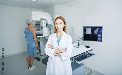 Portrait of mammologist with arms crossed standing in radiology room with imaging machine for breast screening and mature patient. Mammography, mammogram photo