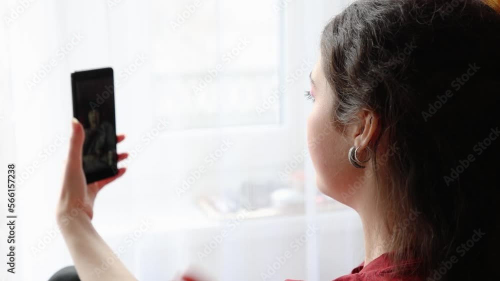 Back view of woman video calls by cellphone. Window in background. Concept of freelance and online communication. Back view of woman video calls by cellphone. Window in background. Concept of freelance and online communication.