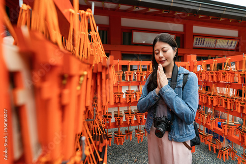 selective focus of pious asian Japanese girl surrounded by red wood plaques ema and making wish with hands together at Fushimi Inari Taisha shrine in Kyoto japan