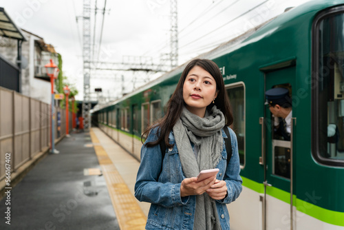 smiling asian Japanese woman visitor consulting guide on phone and looking around on platform of inari station in Kyoto japan on cloudy day. the green train is departing at background