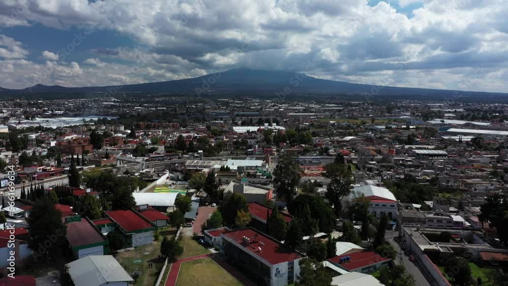 Aerial view of Tlaxcala, Mexico. Camera flies backwards and gains altitude, revealing more of the city, with a dramatic mountain as a backdrop, surrounded by blue skies with scattered white clouds.