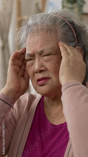 Vertical Screen: shoulder shot of a taiwanese senior lady having sudden headache is rubbing her temples with a grimacing face in the living room at home