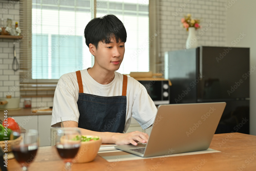 Smiling asian man looking at laptop screen, working online while sitting at wooden table in kitchen Smiling asian man looking at laptop screen, working online while sitting at wooden table in kitchen