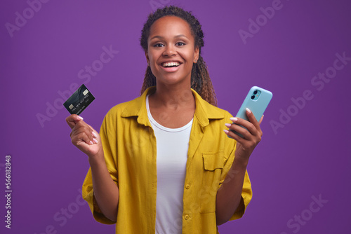 Young positive African American woman with credit card and smartphone enjoy possibility online shopping without leaving your home or favorable prices for buying clothes stands in purple studio