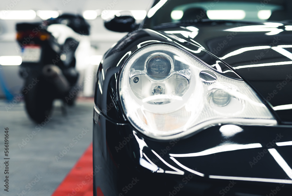 Fototapeta premium Sports car headlamp. Shiny black sports car in garage. Camera focus on the foreground. Blurred white motorcycle in the background. High quality photo