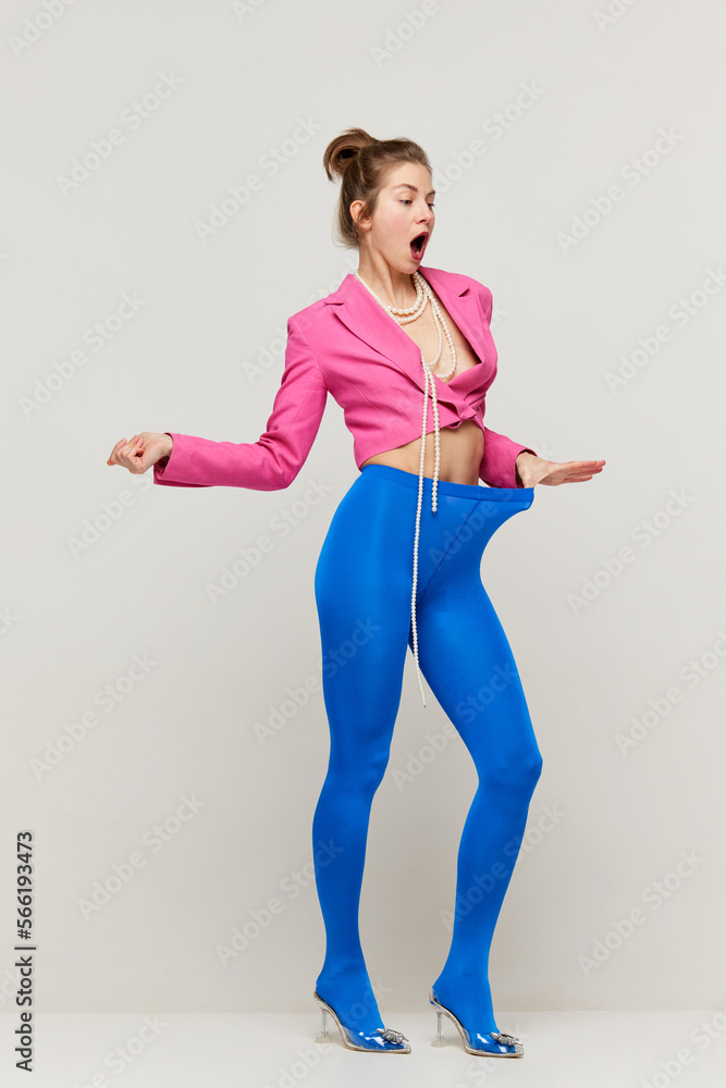 Emotional young stylish girl in blue tights and pink croptop posing, doing strange  poses on floor isolated over grey background. Concept of weird people, art,  fashion Stock Photo