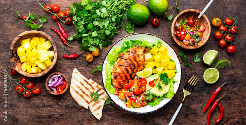 Grilled chicken salad with avocado, mango, tomato salsa, cilantro and lettuce in mexican style, rustic wooden table background, top view
