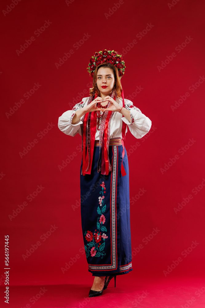 Ukrainian woman in traditional ethnic clothing and floral red wreath on viva magenta studio background with hands showing a heart shape. Ukrainian national embroidered dress call vyshyvanka