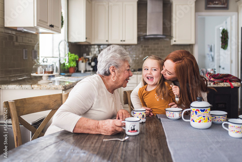 Mother kissing daughter at kitchen table with grandmother looking