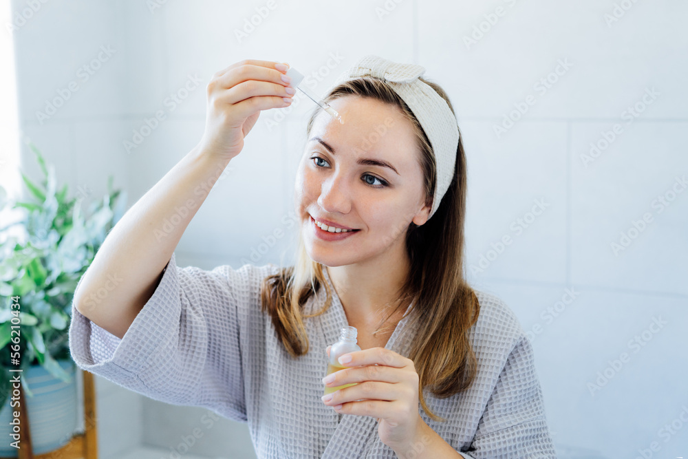 Young woman in bathrobe looking in the mirror, holding dropper with hyaluronic acid serum close to face and smiling. Skin hydrating. Cosmetic spa procedures. Beauty self-care at home. Selective focus Young woman in bathrobe looking in the mirror, holding dropper with hyaluronic acid serum close to face and smiling. Skin hydrating. Cosmetic spa procedures. Beauty self-care at home. Selective focus