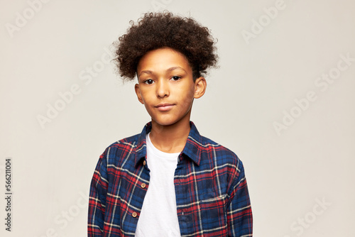 Studio image of smart serious teenage boy with afro hairstyle in plaid shirt posing in gray studio background, looking at camera with no smile. Childhood, youth and teenage maximalism