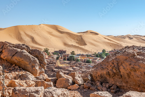Taghit town ksar of Bechar, Algeria Sahara desert. Palm trees and trees, old dry rocks buildings and sand dune with far tiny tourists and a blue clear sky. From Djebel Baroun rocks in foreground. photo