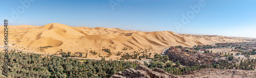 Panorama of Taghit town in Bechar, Algeria Sahara desert. Palm trees oasis, old ksor buildings and sand dunes with a blue clear sky. From Djebel Baroun mountain with unrecognizable tourists far away.