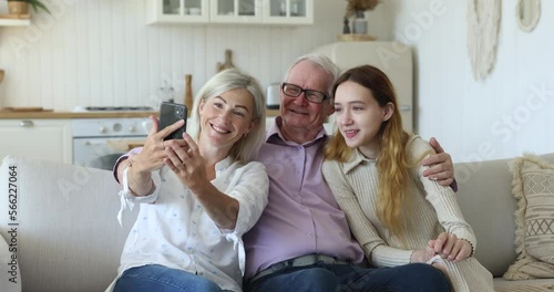 Happy multi generational family spend leisure rest on sofa staring at cellphone screen enjoy new application or take selfie. Grandpa his daughter and pretty teenage 12s granddaughter using modern tech photo