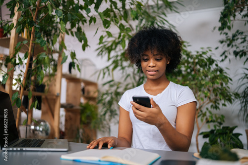 Portrait of focused African-American young woman chatting on digital gadgets online, holding mobile phone, using laptop computer sitting at desk in green home office room with modern biophilia design.