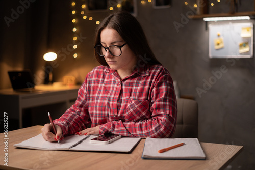 Young woman, teenage girl, student, schoolgirl sits at a desk in a dark dormitory room, writes notes in a notebook and concentrated prepares for the exam.