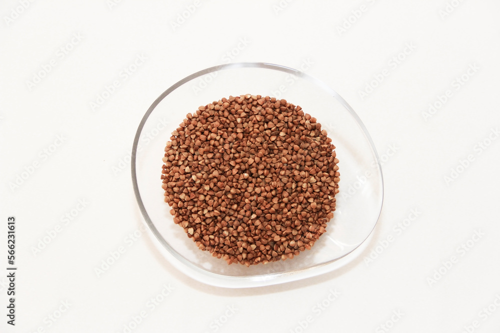 Small grains of natural brown buckwheat in a transparent plate on a light background. Small grains of natural brown buckwheat in a transparent plate on a light background.