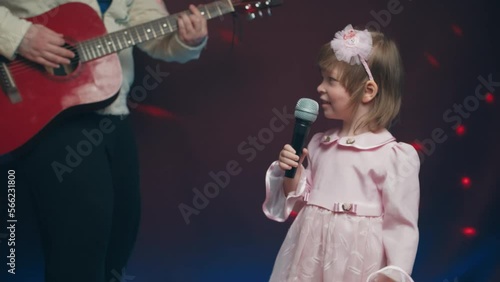 Little girl on stage in vintage dress, she sings into microphone and dances, her father plays acoustic guitar. Color music is shining. Performance on stage. Early development of children
