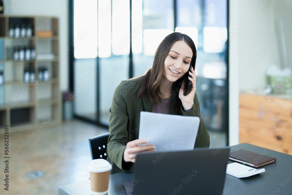 Portrait of a young Asian woman showing a smiling face as she uses his phone, computer and financial documents on her desk in the early morning hours Portrait of a young Asian woman showing a smiling face as she uses his phone, computer and financial documents on her desk in the early morning hours